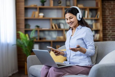 Kulaklık takan ve bilgisayarındaki video görüşmesi sırasında gülümseyen bir kadın, ev ofisinden uzaktan çalışıyor veya rahat bir koltukta otururken internetten öğreniyor.
