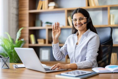 Genç Hintli kadın kameraya gülümsüyor ve kameraya el sallıyor. Video konferansı sırasında kulaklığı açık çalışıyor. Uzaktan çalışma, çevrimiçi eğitim ve sanal iletişimi temsil ediyor.