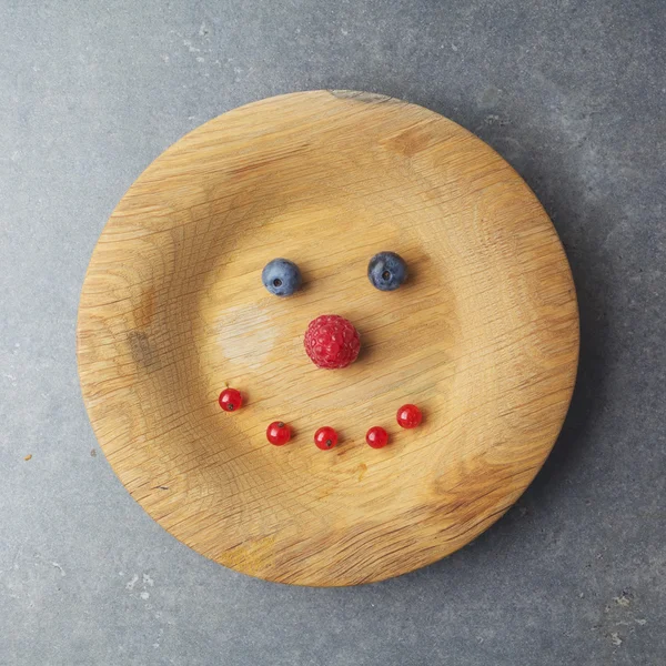 Rosto sorridente em uma placa de madeira com framboesas e mirtilos — Fotografia de Stock