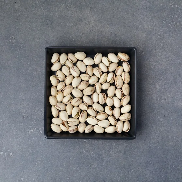 Salted roasted pistachios nuts with shell and salt in black small plate over gray rusty textured background. Top view. Square image — Stock Photo, Image