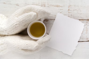 Womans hands in gloves holding a cup of tea with blank card on white wooden table