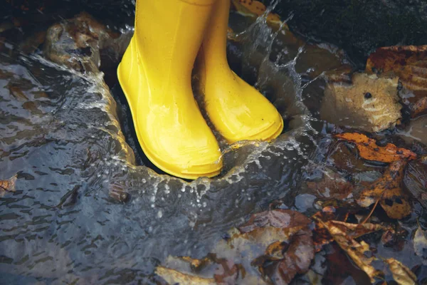 Pies de niño en botas de goma amarillas saltando en charco en el día de otoño. — Foto de Stock