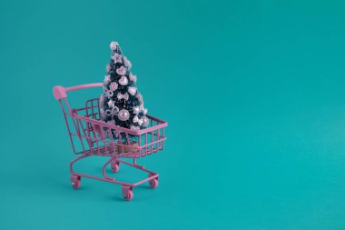 Su rengi arka planda süslü Noel ağacı olan pembe alışveriş arabası..