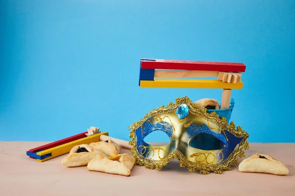 Concepto de celebración Purim con galletas hamantashen, máscara Purim y ruidoso juguete sobre fondo azul — Foto de Stock