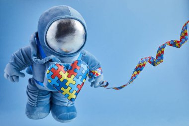 Dünya Otizmi farkındalık günü geçmişi. Mavi pelüş astronot oyuncağı, bulmaca kalpli, otizm sembollü, mavi arka planda