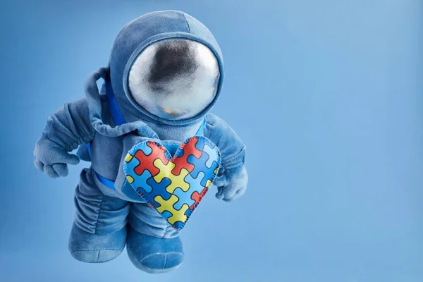 World Autism awareness day background. Blue plush astronaut toy with puzzle heart, autism symbol, on blue background