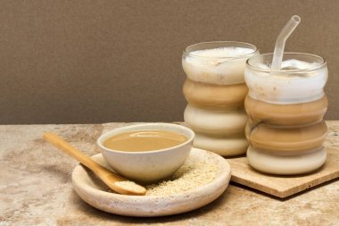 İki bardak katmanlı Tahini Latte kahvesi dokulu bir yüzey üzerindeki susam ezmesinin yanında..