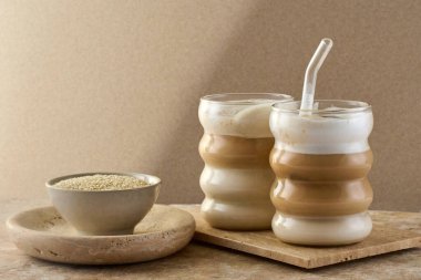 İki bardak katmanlı Tahini Latte kahvesi dokulu bir yüzey üzerindeki susam ezmesinin yanında..