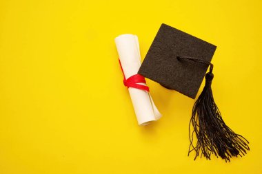 Mezuniyet şapkası ve akademik diploma tomarı sarı arka planda eğitim başarısını simgeliyor