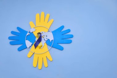 Dünya Down Sendromu Günü kavramı kağıt ellerle çevrili bir kürede mavi ve sarı farkındalık kurdelesini gösteriyor.