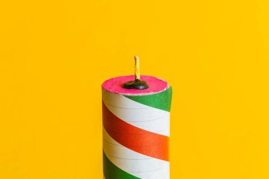 Close-up of a striped firecracker with red, green, and white colors on a vibrant plain yellow background, ready to ignite