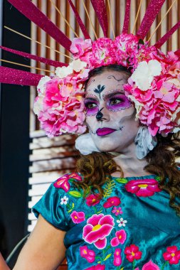Fuşya çiçeği başlıklı ve kafatası makyajlı, geleneksel nakışlı Meksika kıyafetleri giyen genç bir kadının zarif portresi..