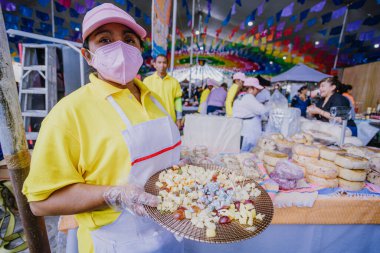 Yiyecek pazarında gülümseyen bir satıcı, güvenlik üniforması giyiyor, tadımlık peynirli bir tabak sunuyor..