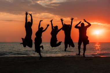 Sahilde sıcak ve renkli bir yaz günbatımında neşeyle atlayan beş kişinin silueti..