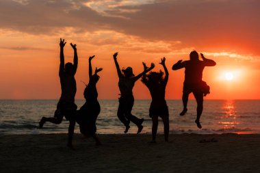 Sahilde sıcak ve renkli bir yaz günbatımında neşeyle atlayan beş kişinin silueti..