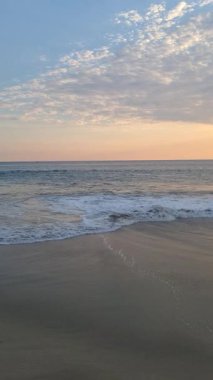Sahil manzarası yumuşak dalgalar kıyıya ulaşıyor mavi gökyüzü altında beyaz bulutlar ve ılık günbatımı tonlarıyla.