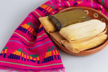 Mısır kabukları ve muz yapraklarına sarılmış bir grup tamale, kil tabakta servis edilir ve renkli zanaatkar tekstil