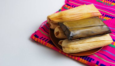 Mısır kabukları ve muz yapraklarına sarılmış bir grup tamale, kil tabakta servis edilir ve renkli zanaatkar tekstil