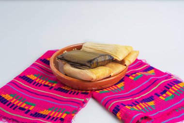 Mısır kabukları ve muz yapraklarına sarılmış bir grup tamale, kil tabakta servis edilir ve renkli zanaatkar tekstil