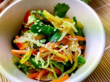 Soğuk sebze salatası ve mantarlı sebze karışımı beyaz bir kasede bambu hasırının üzerinde servis edilir. Havuç, kırmızı, yeşil ve sarı biber, kişniş ve susam tohumları var..