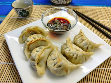Jiaozi adında sirke, sarımsak ve soya soslu Çin usulü kızarmış hamur köftesi. Beyaz kare tabakta ve yanında çubuklarla bambu hasırında servis ediliyor..