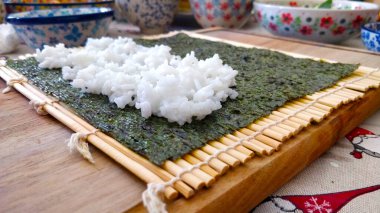 Suşi hazırlarken atılacak adımın ayrıntıları. Pirinç, bambu hasırının üzerindeki yosun kağıdına yayılır. Arka planda seramik bardaklar var..