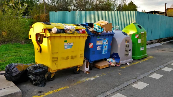 Dustbins for recyclable waste in Prague, Czech Republic. There is bin for paper, plastics, metal, and glass. Behind the garbage bins are trees.