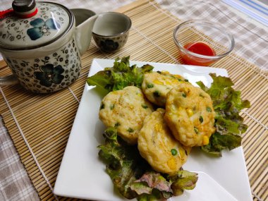Bambu hasırının üzerinde duran beyaz tabakta servis edilen marullu kızarmış karides omleti. Ayrıca acı biber için küçük bir kase çay demliği var..