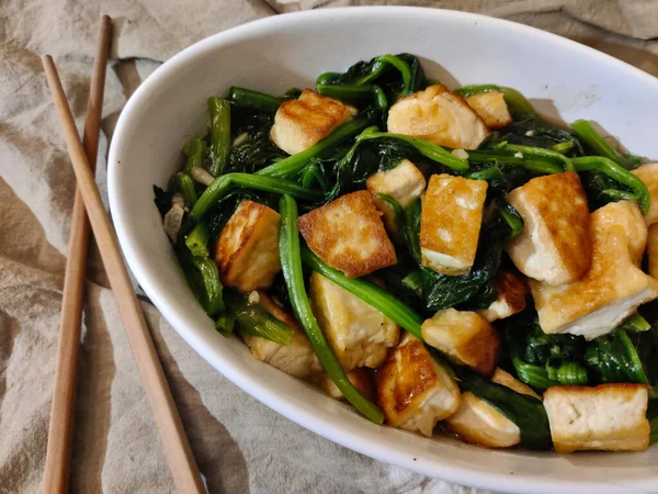 Oval kasede servis edilen haşlanmış pak choi ile kızarmış tofudan yapılmış bir tabak. Kasenin yanında bir çift yemek çubuğu var ve her şey kahverengi masa örtüsünün üzerinde..