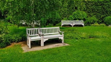 Görüntünün önünde arka planda iki bank olan beyaz bir bank. Resmin her yerinde çimenler var ve koltuğun arkasında da uzun çitler var..