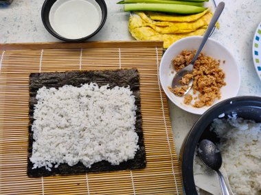 Suşi hazırlama sürecindeki adımlardan biri, beyaz pirincin bambu hasırının üzerinde duran yosunların üzerine yerleştirilmesidir. Doldurmak için kullanılan tüm malzemelerin olduğu kaseler de var..