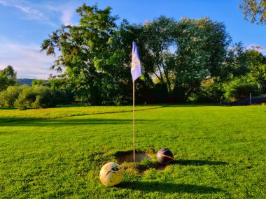 Futbol sahasında bayraklı ve etrafında iki top olan bir delik. Arka planda ağaçlar olan güzel biçilmiş çimlerle oynanır..