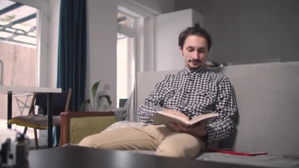 El tipo está leyendo un libro sentado en el sofá — Vídeo de stock