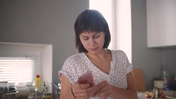 Una chica en la cocina con un teléfono en la mano — Vídeos de Stock