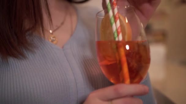 A young brunette with long loose hair drinks a cocktail from a glass with a straw — Wideo stockowe