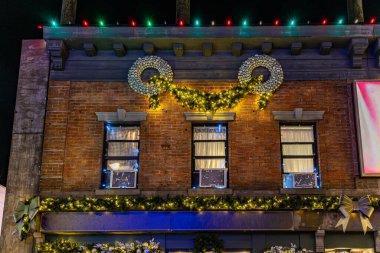 Zarif çelenkler, çelenkler, kurdeleler, fiyonklar ve tatil ışıklarının yer aldığı Noel için dekore edilmiş şenlikli kentsel tuğla bina. Aydınlatılmış pencereler sıcak, kış gecesi atmosferi yaratır. Tatil, şehir hayatı ve mevsimsel kutlamalar için mükemmel bir tema..