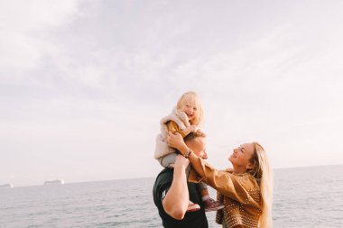 Anne, baba ve küçük kız denizin kenarında gülümsüyorlar. Sarı saçlı, kürklü bir kız çocuğu. Bahar geldi, güneş parlıyor. Yurt dışına seyahat et. Mutlu genç aile. 