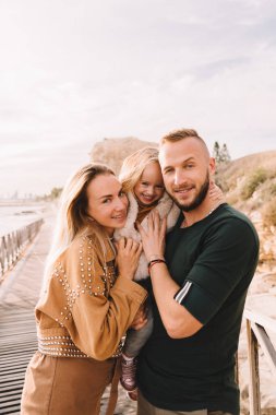 Anne, baba ve küçük kız denizin kenarında gülümsüyorlar. Sarı saçlı, kürklü bir kız çocuğu. Bahar geldi, güneş parlıyor. Yurt dışına seyahat et. Mutlu genç aile. 