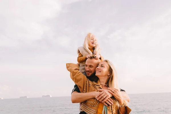 Anne, baba ve küçük kız denizin kenarında gülümsüyorlar. Sarı saçlı, kürklü bir kız çocuğu. Bahar geldi, güneş parlıyor. Yurt dışına seyahat et. Mutlu genç aile. 