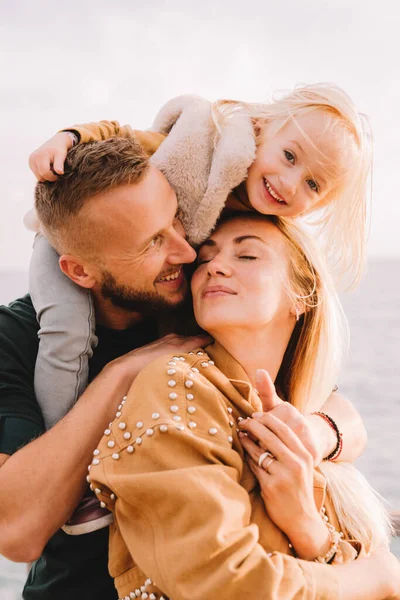 Anne, baba ve küçük kız denizin kenarında gülümsüyorlar. Sarı saçlı, kürklü bir kız çocuğu. Bahar geldi, güneş parlıyor. Yurt dışına seyahat et. Mutlu genç aile. 