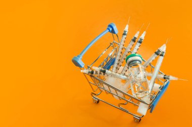 Turuncu arka planda şırınga ve ampul bulunan süpermarket arabası