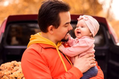 Sonbahar parkında, çiftlikte ya da ticaret fuarında kucağında pembe şapkalı küçük kız çocuğunu tutan bir baba. Arka planda çiçeklerle dolu bir kamyonet yatağı. Baba ve küçük çocuk sonbahar aile hafta sonunu dışarıda geçiriyorlar.