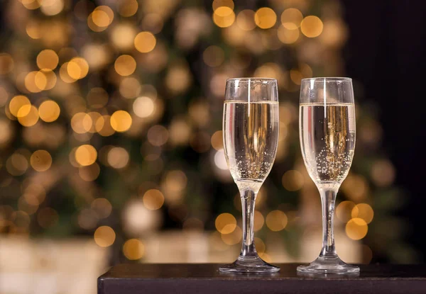Two flute glasses with sparkling drink (champagne) standing on dark wooden table. Blurred decorated Christmas (New year) fir tree in background. Romantic winter holidays concept. Copy space