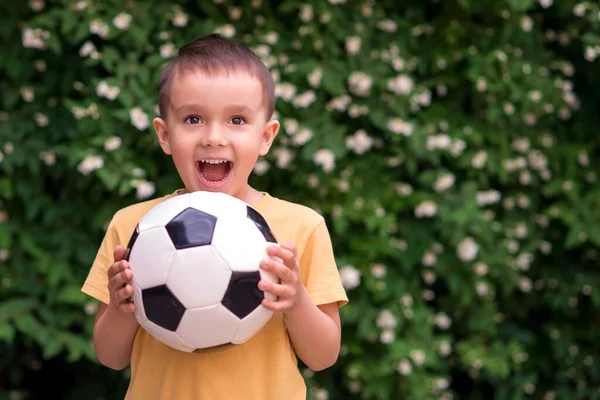 興奮した小さな幼児の少年は屋外でサッカーを保持しています 緑の葉と花の後ろ 子供はボールをキャッチし 幸せと感情を叫んでいました 積極的な子供時代とスポーツの情熱の概念 スペースのコピー — ストック写真