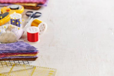 Cetvel, kesme hasırı, çok renkli kumaş yığını, iğnelik, iplik makaraları, makas, döner kesici, beyaz bir yüzeyde kumaş yığını. Metin için boşluk