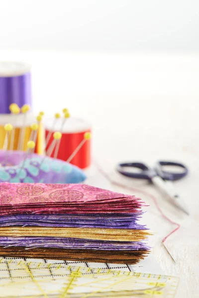 Ruler, cutting mat, stack of multi-colored pieces of fabric, pincushion, spools of thread, scissors, needle and thread on a white surface. Space for text