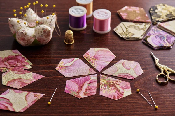 Pentagonal pieces of fabric lying in the shape of a flower, stacks of multi-colored pieces of fabric, sewing accessories on a desk