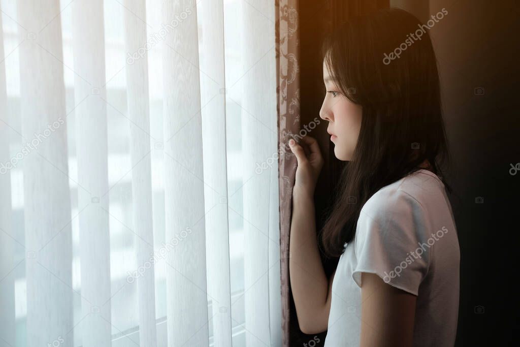 A depressed Asian woman stands at the window.