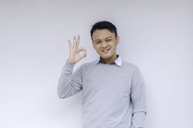Genç Asyalı adam mutlu gülümseyen yüzü ve baş parmağı olan gri bir gömlek giyer..