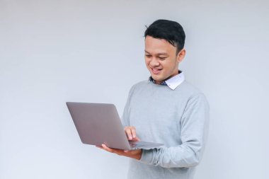 Genç Asyalı adam ayakta durup dizüstü bilgisayarla çalışırken gülümsüyor. Gri gömlekli Endonezyalı adam.